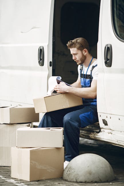 A man with a beard, wearing a white and blue striped t-shirt and navy work overalls, sits on the edge of a white moving van with sliding doors open, holding a clipboard and pen. Surrounding him are several cardboard boxes of varying sizes, some stacked on the ground nearby, ready for home relocation or furniture transport. The scene is set outdoors on a paved surface with a concrete bollard visible in the foreground. Inside the van, packing materials such as white packing paper or bubble wrap can be seen, indicating a packing and moving process. The lighting is natural, suggesting daytime, and the overall environment reflects a professional moving operation, with the man representing a mover from Man With a Van Epsom, a company offering removals services as highlighted on their website page about avoiding hidden fees with quotes.