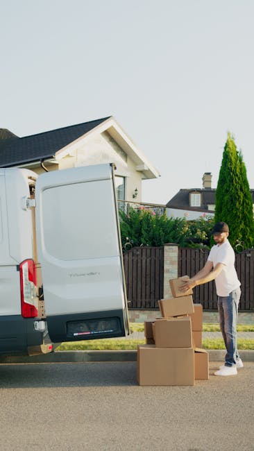 A man in casual clothing and glasses is standing beside an open rear door of a white moving van parked on a residential street in Epsom, surrounded by green trees and a wooden fence. He is handling ca