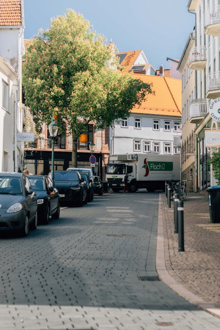 A wide view of a narrow street in Epsom, KT17, with parked cars along both sides and a large green tree in the centre. Multi-storey white buildings with orange-tiled roofs line the street, and a white moving truck with the logo 'Flachas' is partially visible near a building. The street surface consists of cobblestone paving, and there are street lamps and low black bollards protecting the pavement edge. The environment is bright with natural sunlight, indicating daytime. This scene captures a typical urban setting suitable for house removals or relocation services, with no visible active moving activities but highlighting the area where professional furniture transport and packing and moving operations may take place, as seen with the parked vehicles and urban surroundings. Man With a Van Epsom occasionally operates in such environments for home relocations and furniture transport services.