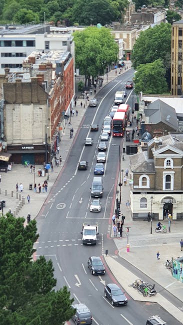 An aerial view of a busy street in Epsom, KT17, showing parked cars along the curb and moving vehicles including vans and cars in transit, with pedestrians walking on sidewalks. The scene includes a row of multi-storey and single-storey buildings, some with brick facades, and a prominent tree-lined section in the background. At street level, bicycles are parked at a bike rack near a small building. The roadway features clear lane markings, and the area is illuminated by natural daylight, suggesting a calm, weekday environment. This visual captures an urban setting involved in routine traffic movement and pedestrian activity, relevant to house removals and urban moving services such as those offered by Man With a Van Epsom, specializing in furniture transport and home relocation logistics.