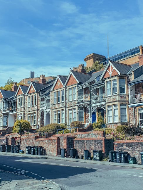 Photograph of a row of Victorian-style terraced houses on Guildford Road Epsom, with distinctive gabled roofs, ornate white iron balconies, and large bay windows. The houses are built from brick and feature decorative trim, set on a raised platform accessible by steps. In front, there is a pavement with a line of black and green wheelie bins, and shrubs or small trees planted along the front gardens. Above the buildings, the sky is partly cloudy with patches of blue visible. This residential setting reflects typical home relocation surroundings in Epsom, suitable for house removals and furniture transport services by Man With a Van Epsom, especially for properties with narrow access points.