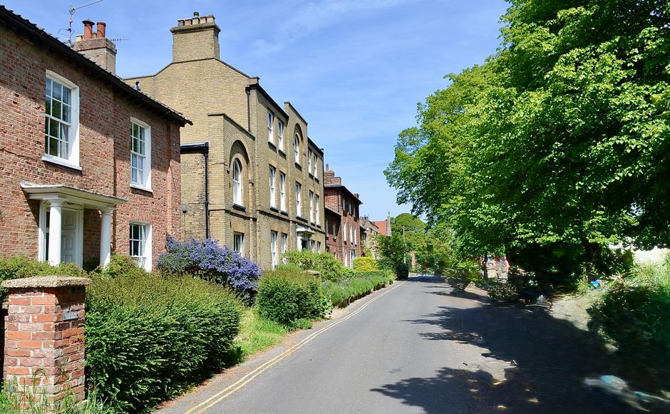 A quiet residential street during daytime with a row of traditional brick houses featuring white window frames and small front gardens, some with purple and green shrubbery. The pavement runs alongside the houses, with a narrow road in the foreground. On the right side, a large, leafy tree with bright green foliage provides shade over part of the pavement and some parked vehicles. The sky above is clear and blue. This setting illustrates a typical environment where home relocation and furniture transport may occur, with the street’s narrow access and greenery highlighting the challenges of moving in such areas. Man With a Van Epsom, a professional house removals company, operates in similar settings facilitating packing, loading, and home relocation services amidst these residential surroundings.
