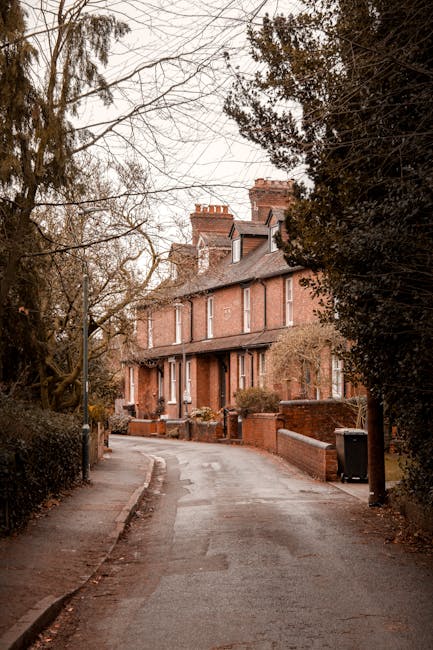 Photograph of a row of Victorian-style terraced houses on Guildford Road Epsom, with distinctive gabled roofs, ornate white iron balconies, and large bay windows. The houses are built from brick and feature decorative trim, set on a raised platform accessible by steps. In front, there is a pavement with a line of black and green wheelie bins, and shrubs or small trees planted along the front gardens. Above the buildings, the sky is partly cloudy with patches of blue visible. This residential setting reflects typical home relocation surroundings in Epsom, suitable for house removals and furniture transport services by Man With a Van Epsom, especially for properties with narrow access points.