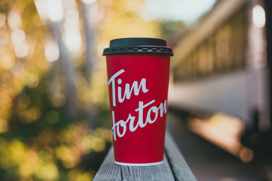 A close-up of a red disposable coffee cup with a black plastic lid, placed on a wooden surface outdoors during daylight. The cup features the white text 'Tim Horton' written in a casual, cursive font. The background showcases a blurred scene with trees and foliage, suggesting an autumn setting with warm, golden tones. To the right of the cup, partial elements of a railway or train station platform are visible, including a metal railing and a train car in the distance, indicating an outdoor transportation environment. The image contextually relates to house removals and moving services by highlighting potential travel or transit during a home relocation, with the focus on travel essentials like beverages and transport infrastructure, associated with Man With a Van Epsom's services in the Horton Lane area near Horton Hospital, EPSOM.